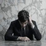 A young man in a suit sits at a table, looking down with a distressed expression, representing the need for mindset mapping to overcome limiting beliefs.